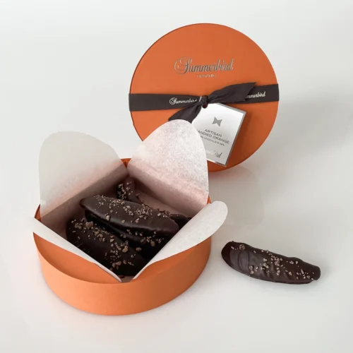 En rund orange gaveæske med et mørkt bånd indeholder Summerbird Artisan Kandiserede Orangestænger, 120 g, chokoladeovertrukne kandiserede appelsinskaller på hvidt pergamentpapir. Et stykke chokolade ligger uden for æsken.